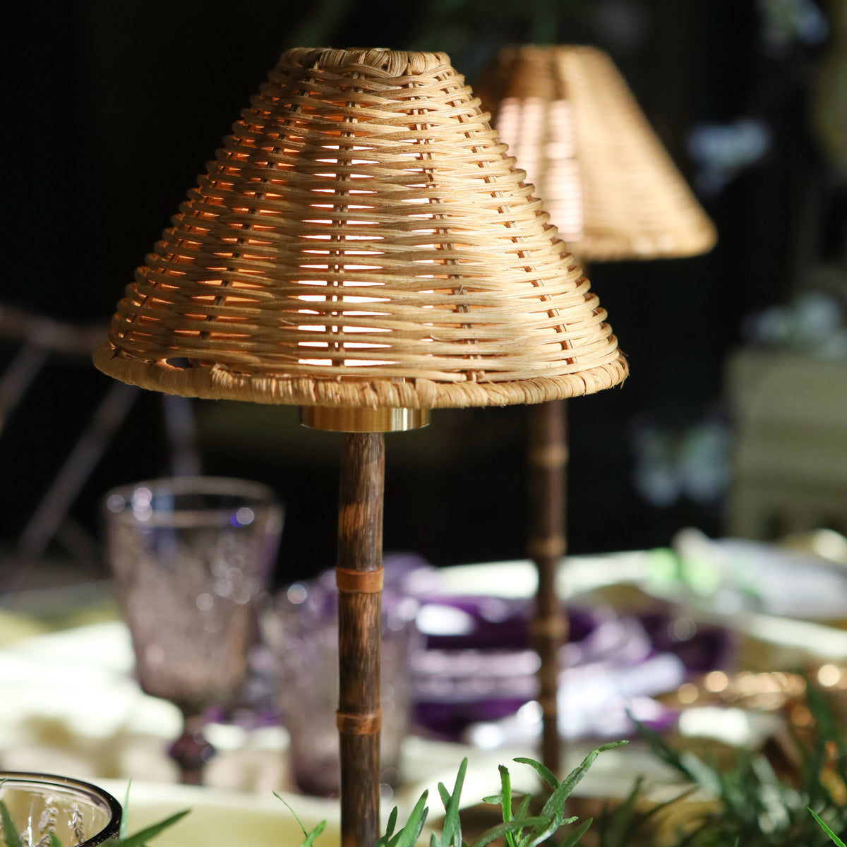 A small table lamp with a woven natural rattan shade and a dark bamboo-style stem, shown in a styled dining or event setting with blurred glassware and greenery in the background.