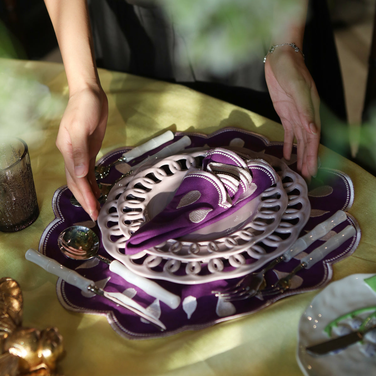 A close-up of a person's hands arranging a decorative purple and silver place setting on a yellow tablecloth. The set includes a scalloped purple charger plate, a silver ornate plate, matching purple and silver cutlery, and white candle-style holders.