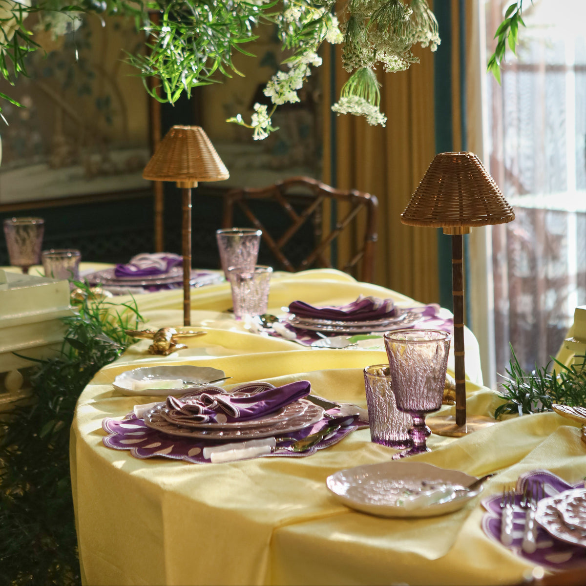 An elegant table setting draped in a pale yellow tablecloth, decorated with purple napkins, matching purple glassware, and woven table lamps, under a lush hanging floral and greenery display.