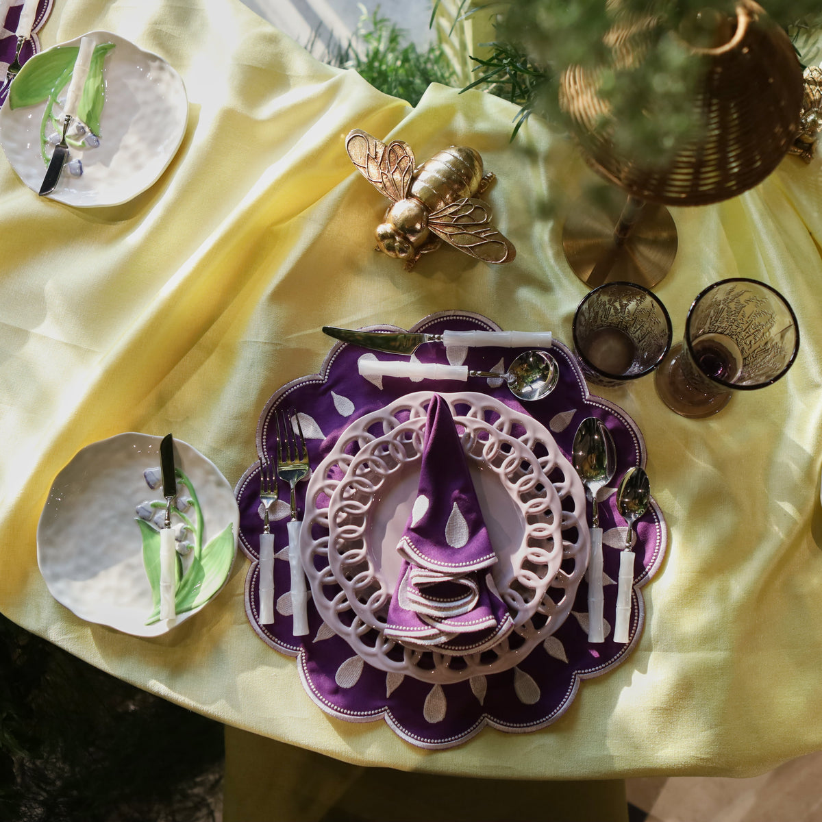 An overhead view of a styled table setting featuring purple and white patterned plates, silver flatware, two dark amber glasses, and a central decorative gold bee figurine, all arranged on a draped pale yellow fabric.