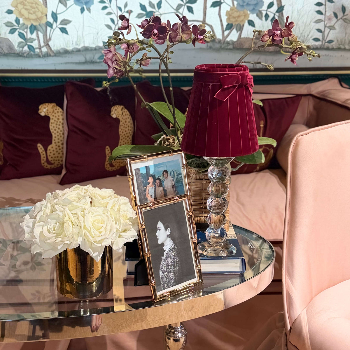 A styled interior scene with a round glass-top table holding a gold vase of white roses, framed photos, and a deep burgundy pleated lampshade on a decorative pedestal. Behind the table is a blush pink sofa with burgundy pillows, and a pale pink upholstered chair is visible in the foreground.