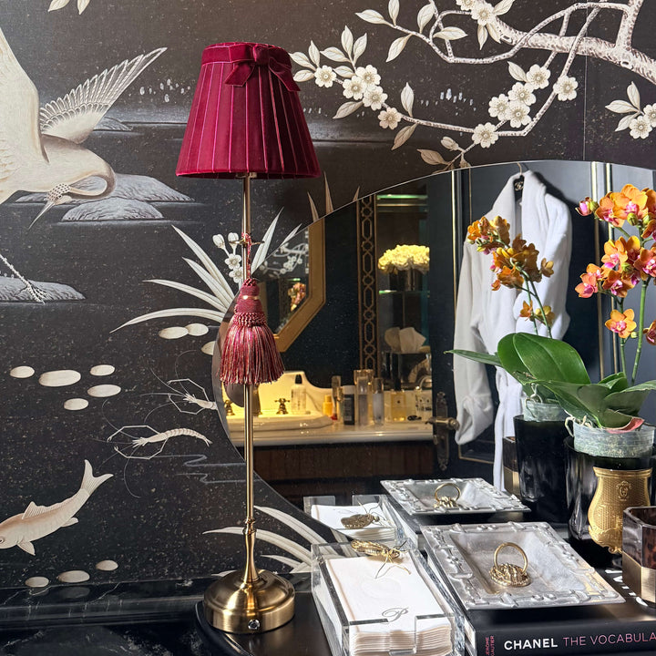 A luxury bathroom vanity setup featuring a dark brown wallpaper with white floral branches and birds, a deep magenta pleated lampshade on a gold stand, and a black vanity table with a Chanel book and jewelry boxes.