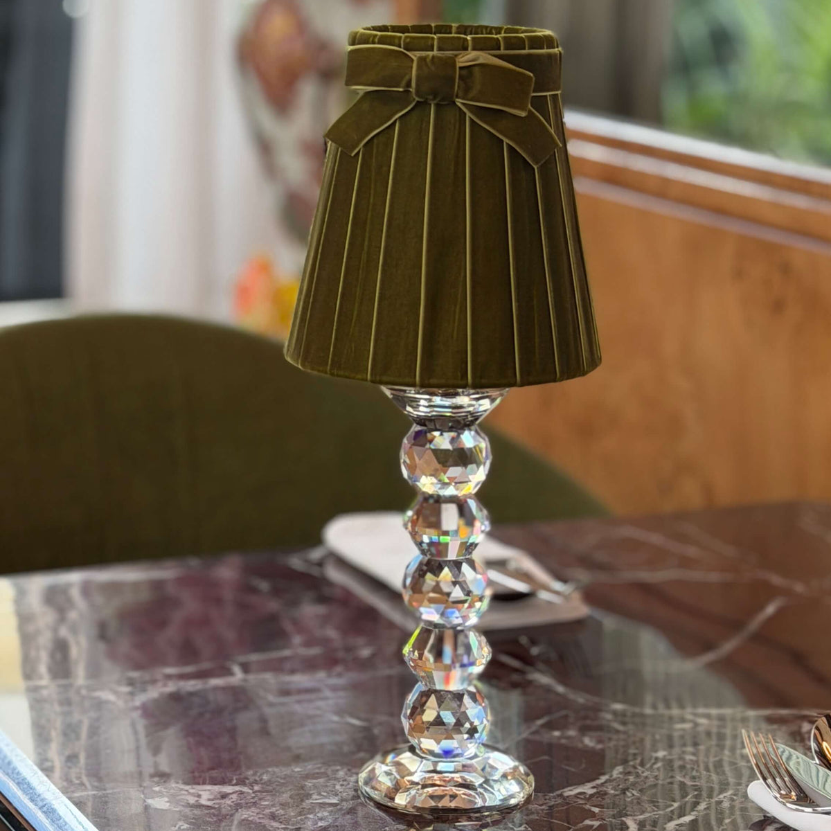 A small table lamp with a pleated olive green fabric shade and a clear faceted glass pedestal base, set on a dark marble tabletop in a softly blurred indoor setting.