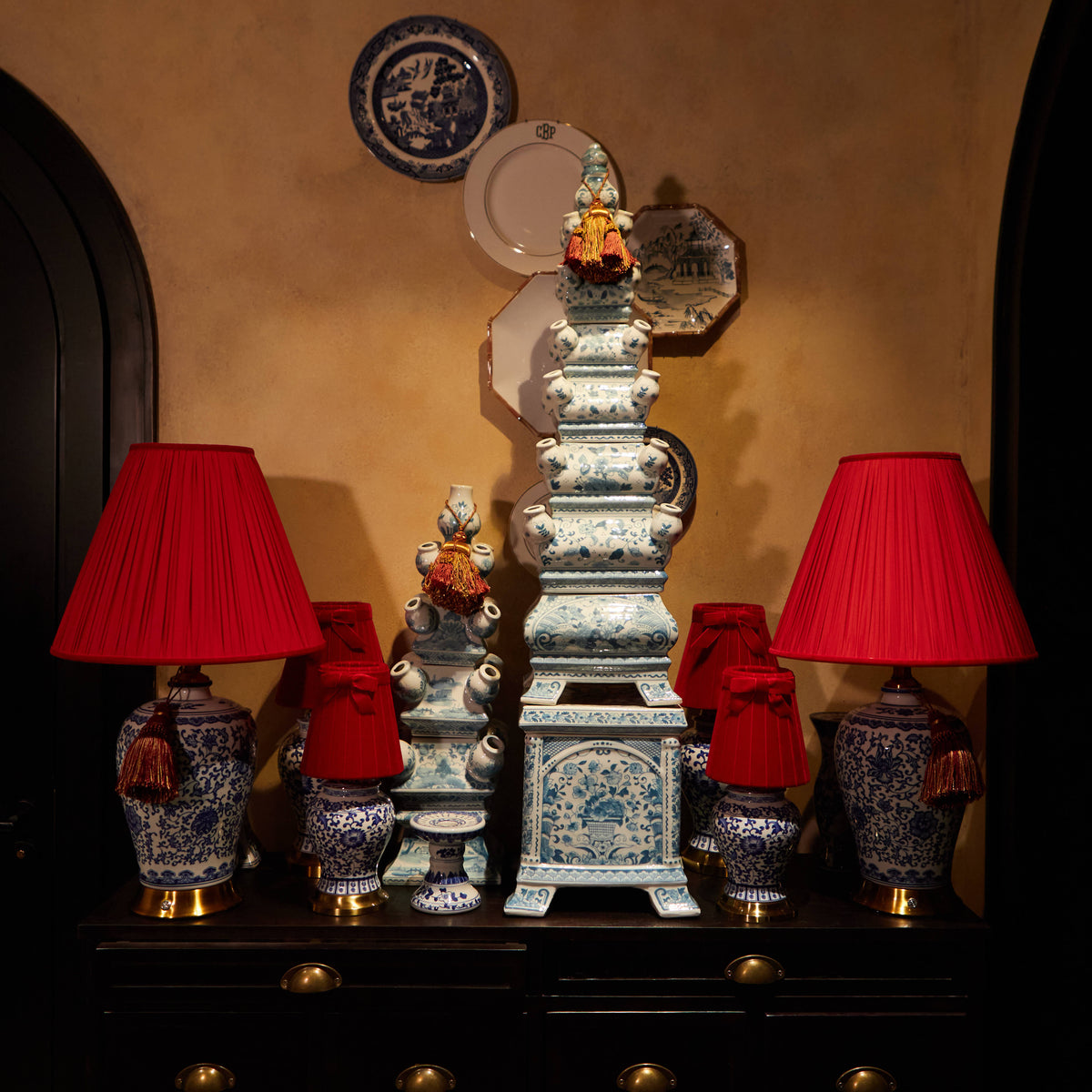 A decorative display featuring a tall blue-and-white porcelain pagoda-style tower centerpiece, flanked by two matching blue-and-white ceramic lamps with bright red fabric shades, set on a dark wooden cabinet against a warm beige wall.