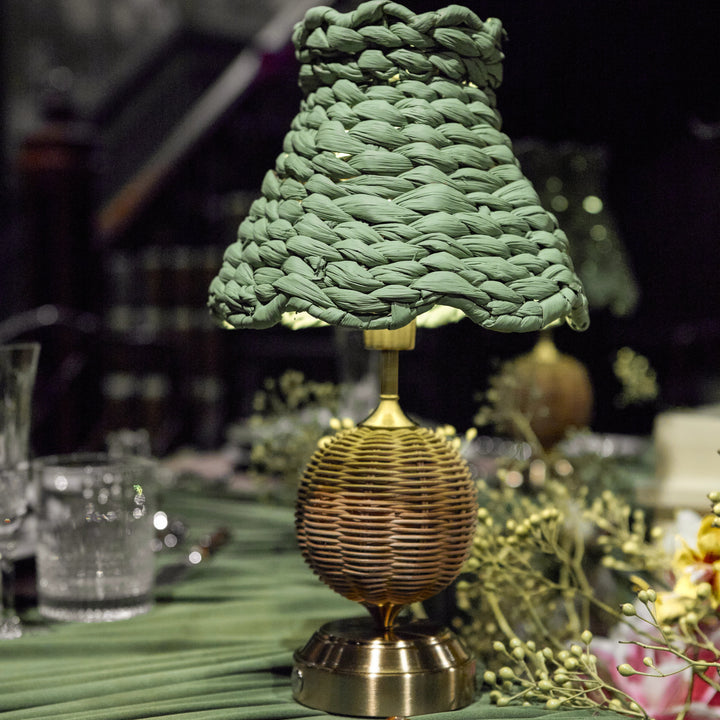 A decorative table lamp with a textured olive green woven shade and a brass ribbed base, set on a green tablecloth with plates and cutlery in a styled dining setting.