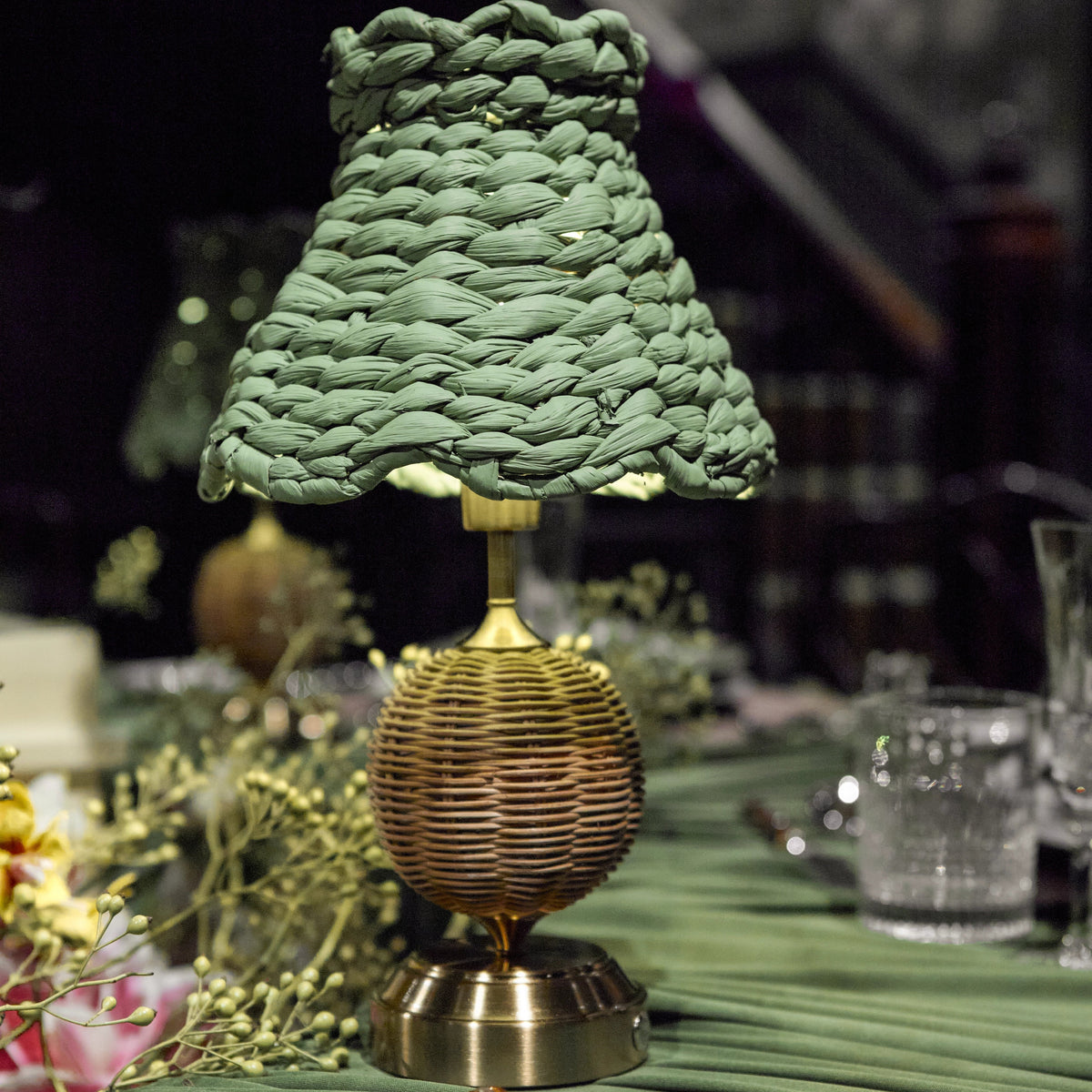 A decorative table lamp with a textured olive green woven shade and a brass ribbed base, set on a green tablecloth with plates and cutlery in a styled dining setting.