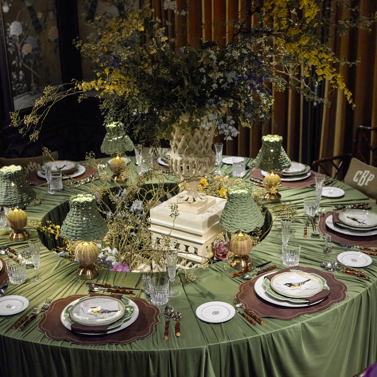 An elegantly set round dining table draped in an olive green tablecloth, decorated with a large floral centerpiece, green textured lamps, white plates, and gold-toned cutlery in an indoor event space.