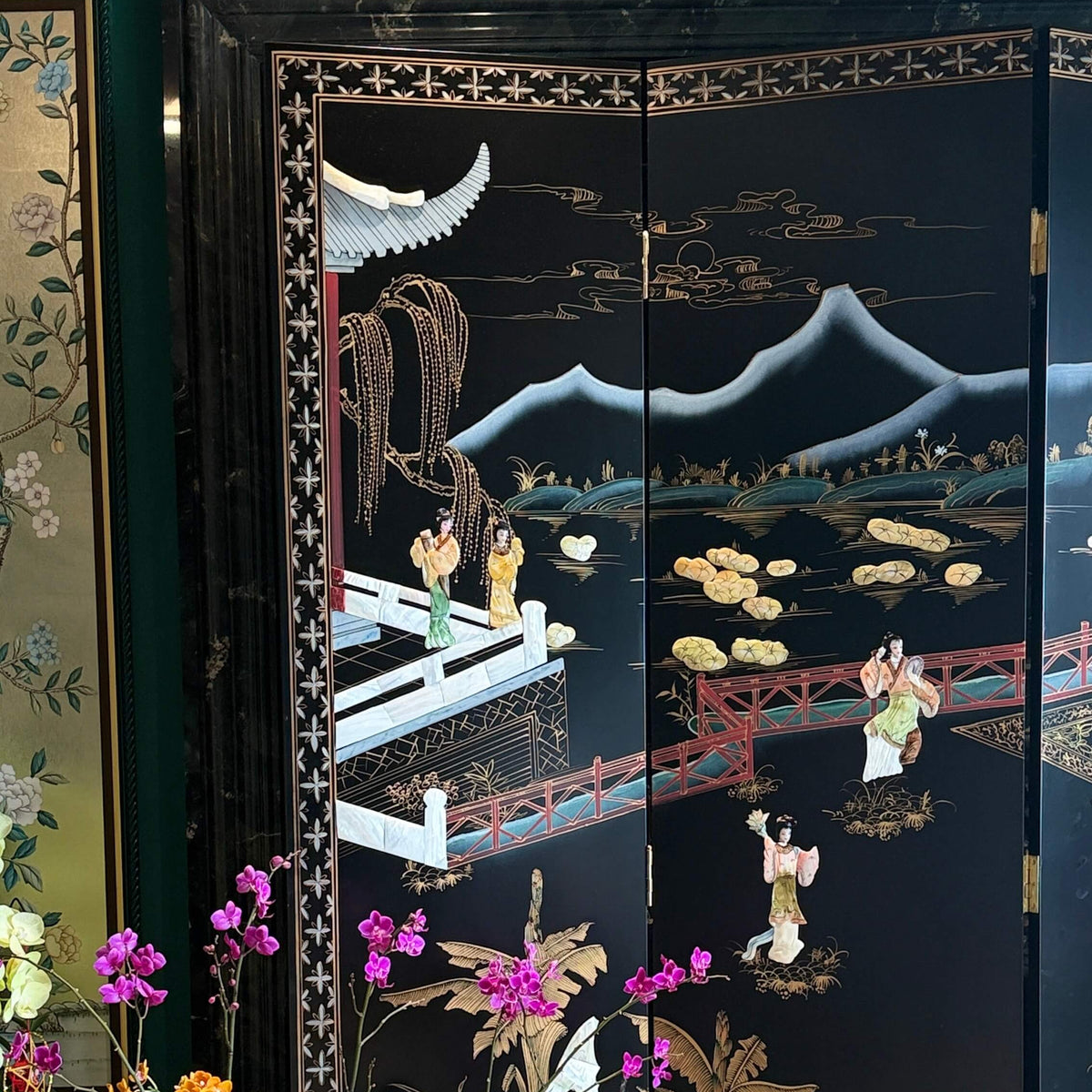 A black lacquered folding screen with intricate hand-painted and inlaid scenes of figures, mountains, boats, and water lilies, styled in a room next to a green glass vase with flowers.