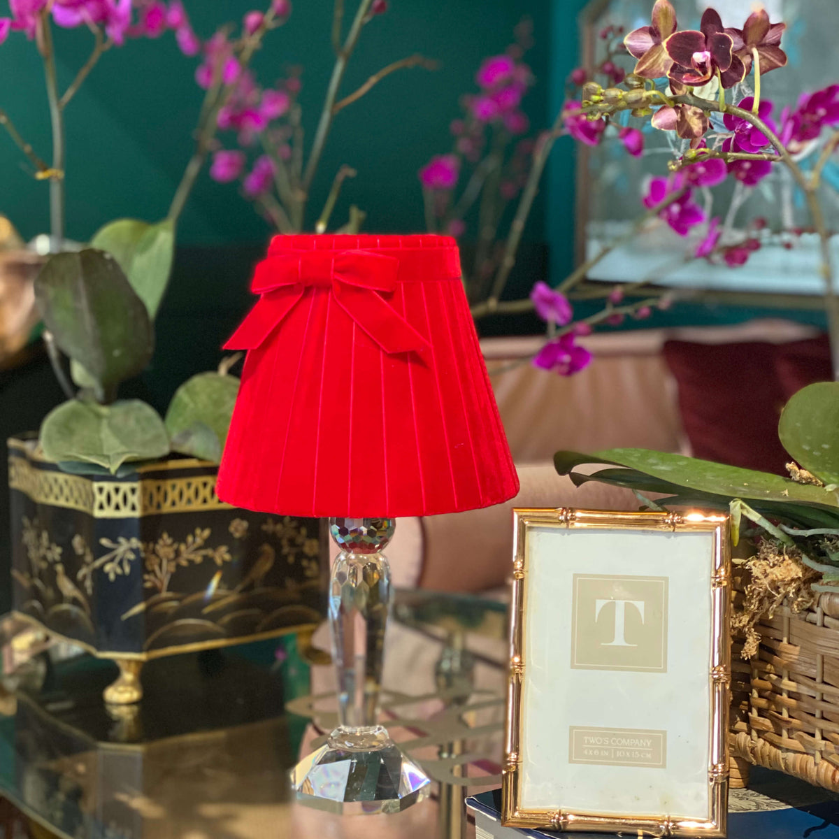 A small pleated red fabric table lamp with a matching red bow on a clear glass stem base, styled on a reflective glass table next to a gold picture frame and books in an indoor setting with purple orchids.