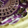Purple embroidered table linens with scalloped edges, a pale pink ceramic charger plate, and silver spoons with white handles arranged on a yellow tablecloth.