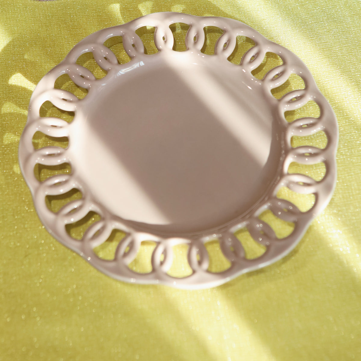 A close-up of a decorative ceramic plate with an intricate scalloped cutout rim, set on a yellow tablecloth alongside other matching tableware.