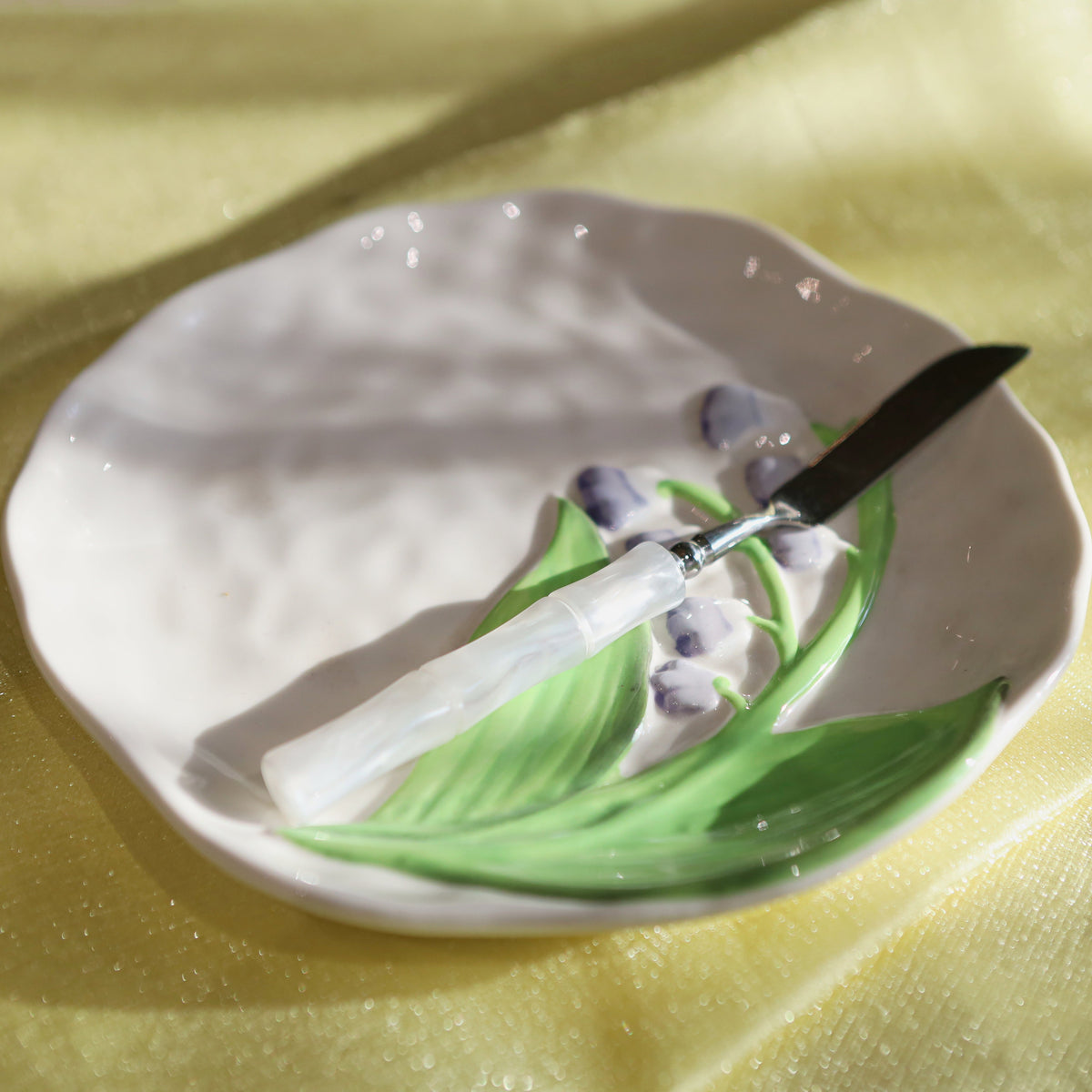 A ceramic plate with a hand-painted lily of the valley design, holding a white-handled knife with a black blade, set against a draped yellow fabric background.