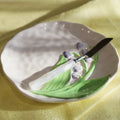 A ceramic plate with a hand-painted lily of the valley design, holding a white-handled knife with a black blade, set against a draped yellow fabric background.