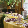 An elegant table setting draped in a pale yellow tablecloth, decorated with purple napkins, matching purple glassware, and woven table lamps, under a lush hanging floral and greenery display.