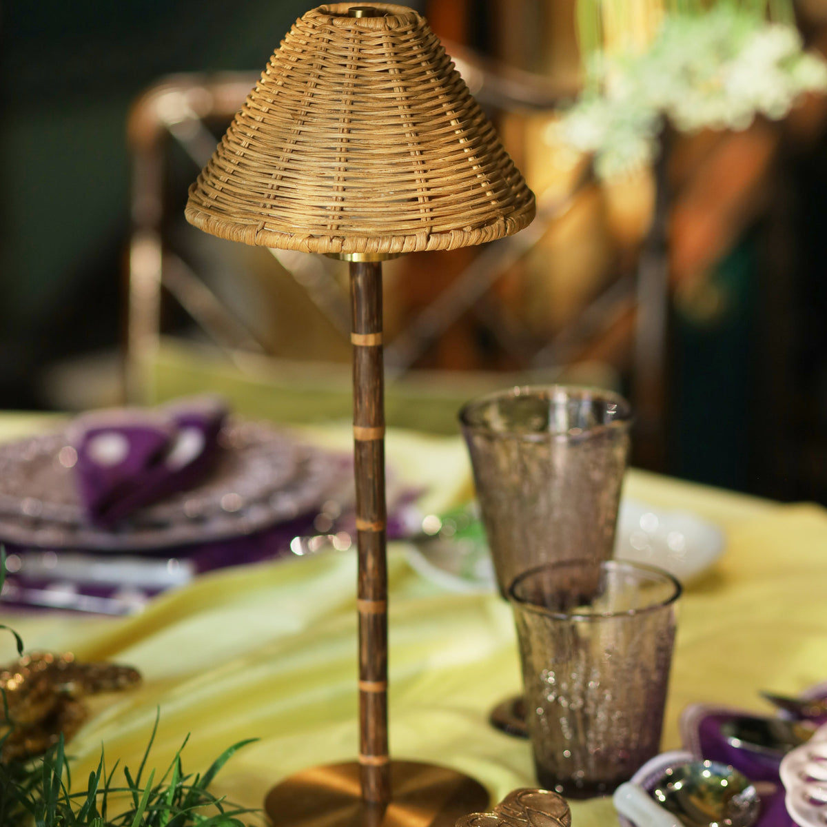 A small table lamp with a woven wicker shade and a metallic bronze stem and base, styled on a yellow tablecloth with purple plates and gold decorative accents.