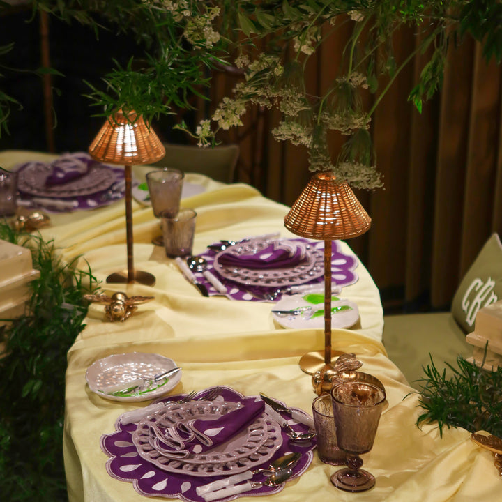 An elegant table setting draped in a cream satin tablecloth, featuring purple and white patterned plates, matching napkins, amber glass goblets, and woven copper table lamps, accented with green foliage.