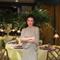 A woman with dark hair tied back stands with arms crossed in an elegant dining room setting. She is wearing a light sage green long-sleeve dress.