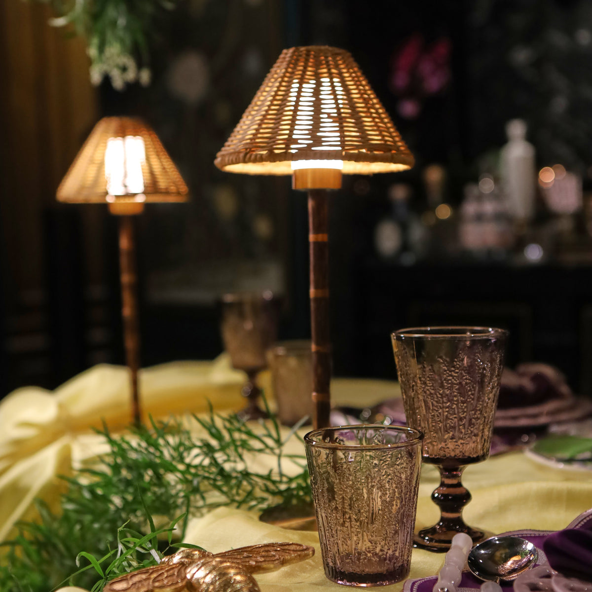 A styled table setting with a pale yellow tablecloth, woven lampshades on two table lamps, amber glass goblets, greenery, and a gold decorative object in a dimly lit indoor environment.
