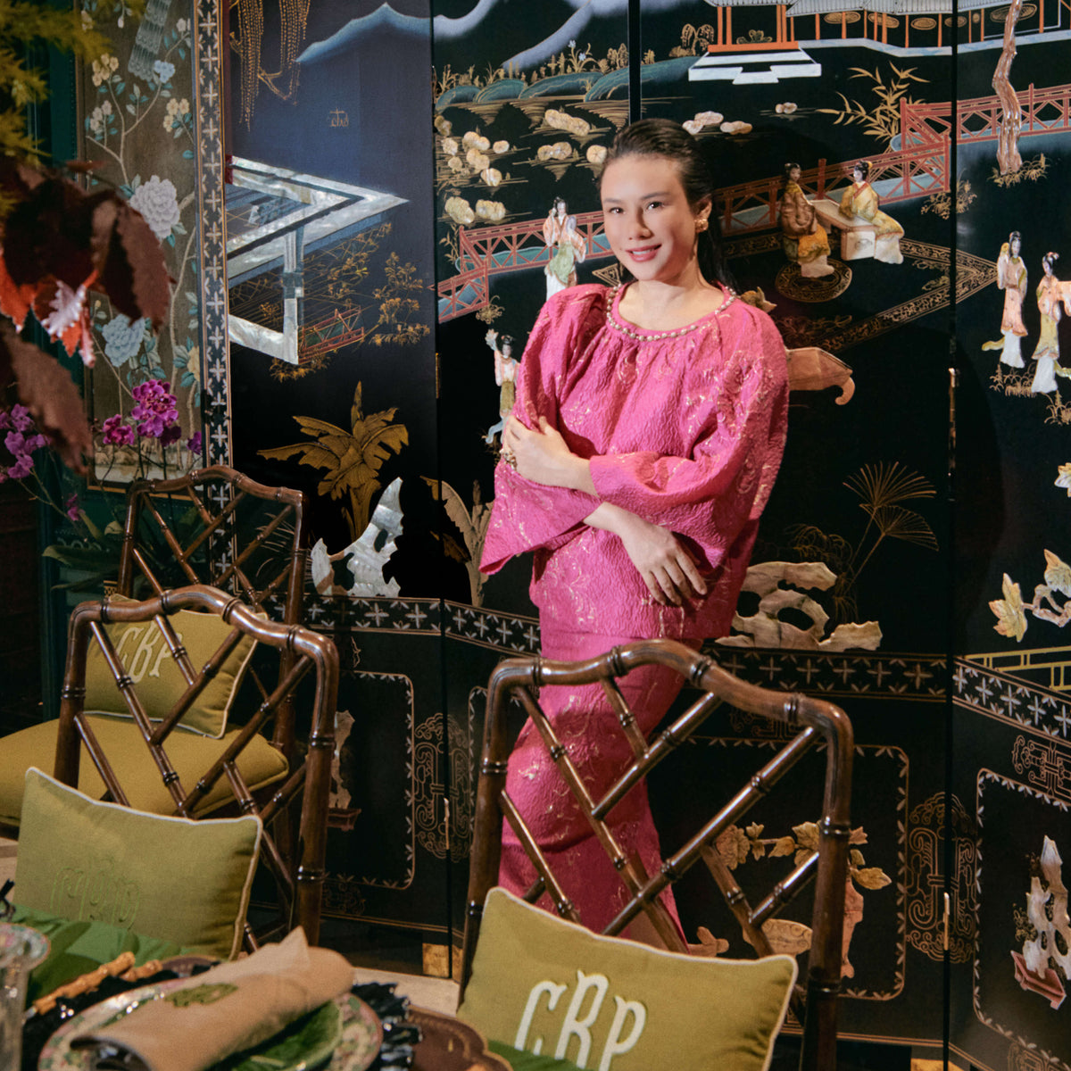 A woman wearing a shiny pink traditional-style dress stands in a decorated interior in front of a black lacquered folding screen with gold and white landscape artwork. A dining table with green tablecloth, plates, and chairs is visible in the foreground.