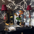 A dark, elegant interior scene featuring a black console table with gold hardware, a large mirror, two red lampshades, and a black wallpaper with white floral branch patterns. A Chanel book is visible on the table.