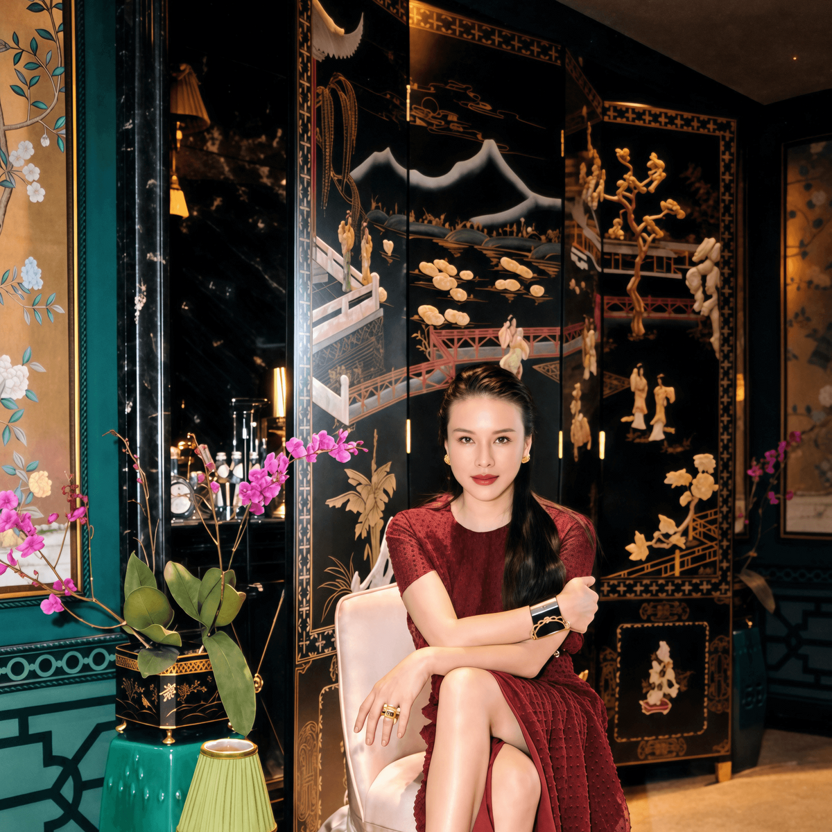 A woman sits on a draped white chair in an ornate interior, wearing a floor-length burgundy dress with short sleeves and a draped skirt. The background features a black lacquered folding screen with gold and white floral and architectural details, alongside a green side table with a lamp and pink flowers.