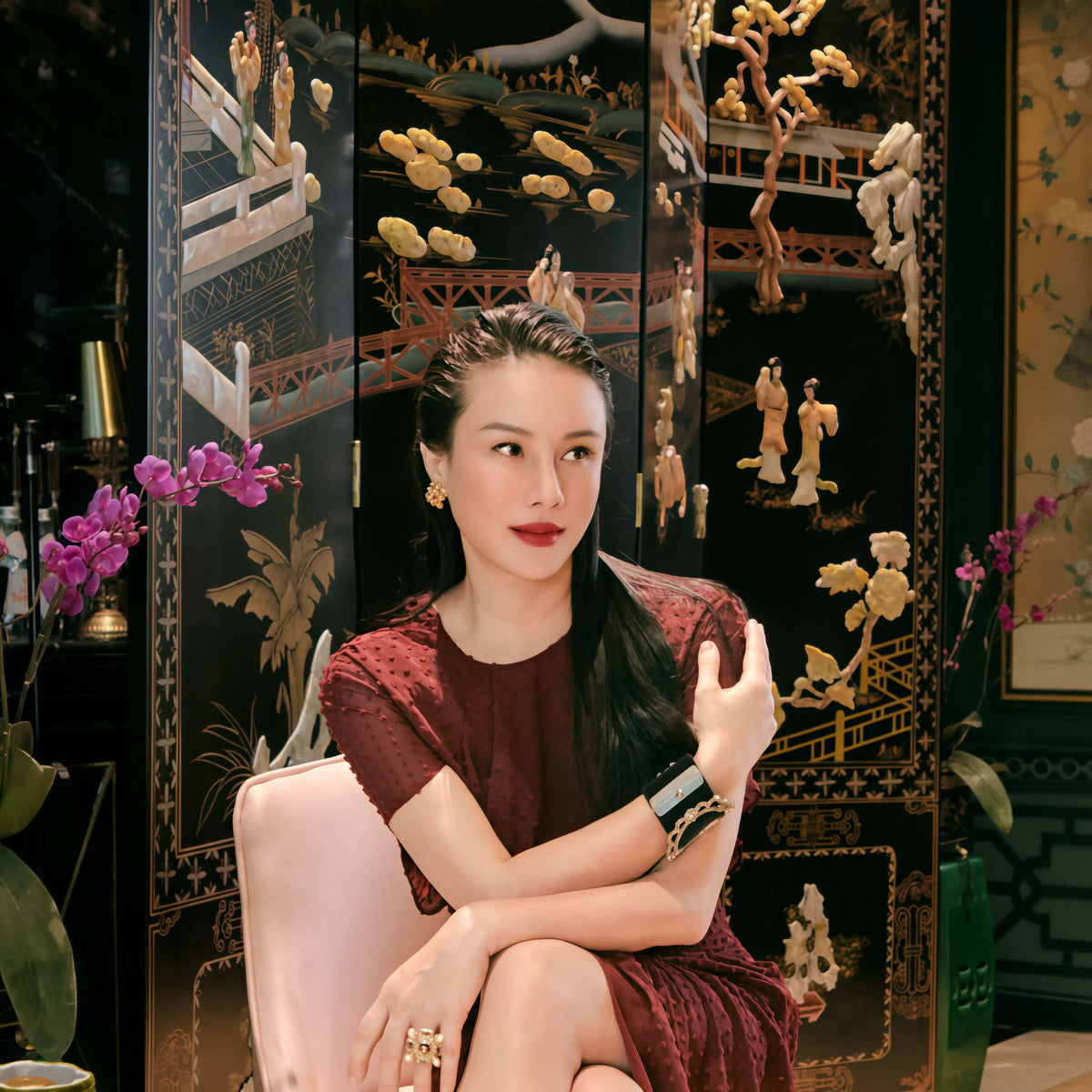 A woman sits in a pale pink armchair wearing a dark burgundy textured dress, posed in front of an ornate black lacquered folding screen with gold and white decorative figures.