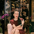 A woman sits in a pale pink armchair wearing a dark burgundy textured dress, posed in front of an ornate black lacquered folding screen with gold and white decorative figures.