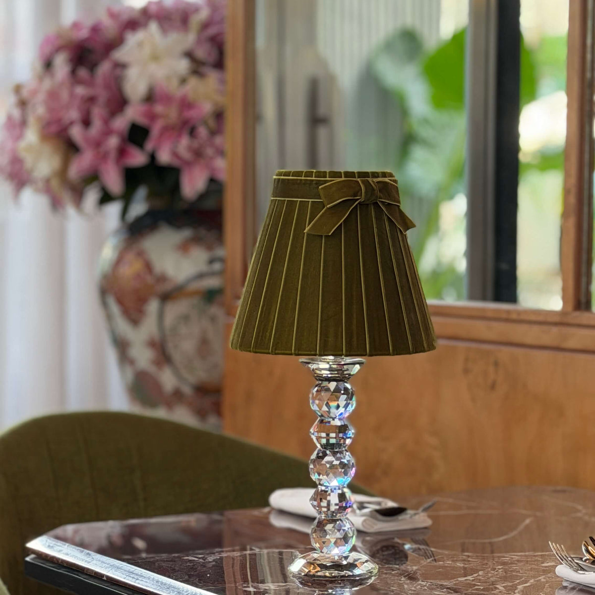A table lamp with a pleated olive green fabric shade and a clear faceted crystal stem, displayed on a polished marble-top table in a dining or living room setting.