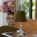 A table lamp with a pleated olive green fabric shade and a clear faceted crystal stem, displayed on a polished marble-top table in a dining or living room setting.