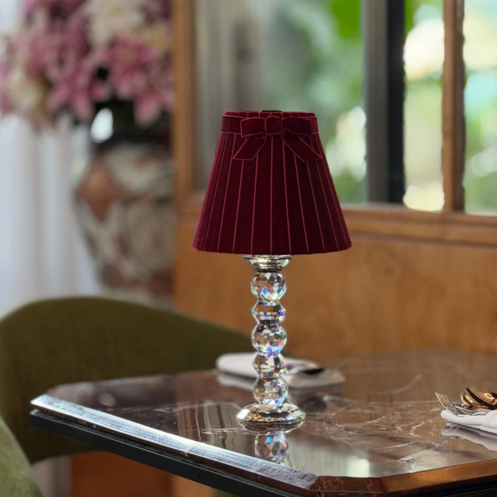 A table lamp with a deep burgundy velvet shade and a clear faceted crystal column base, set on a dark marble-topped table in a dining room setting.