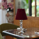A table lamp with a deep burgundy velvet shade and a clear faceted crystal column base, set on a dark marble-topped table in a dining room setting.