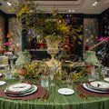 An elegant round dining table set in a richly decorated room, featuring a sage green tablecloth, matching green table lamps, white plates with gold detailing, brown placemats, and a large central floral arrangement in a cream-colored urn.