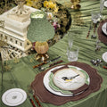 An elegant table setting with olive green table linens, matching textured green lampshades, brown scalloped placemats, white plates with bird illustrations, and decorative glassware.