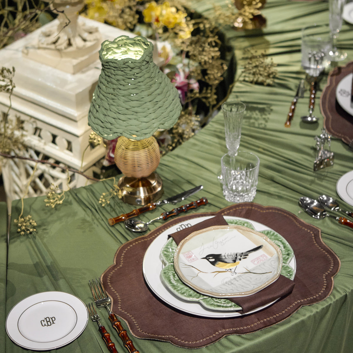 An elegant table setting with olive green table linens, matching textured green lampshades, brown scalloped placemats, white plates with bird illustrations, and decorative glassware.