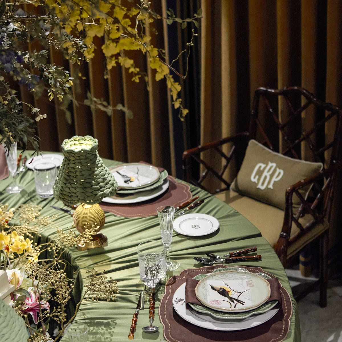 An elegantly styled round dining table draped in olive green fabric, set with white plates, brown placemats, crystal glasses, and a decorative green textured vase. A wooden armchair with a beige cushion marked 'GBP' is visible against a backdrop of brown curtains and yellow foliage.