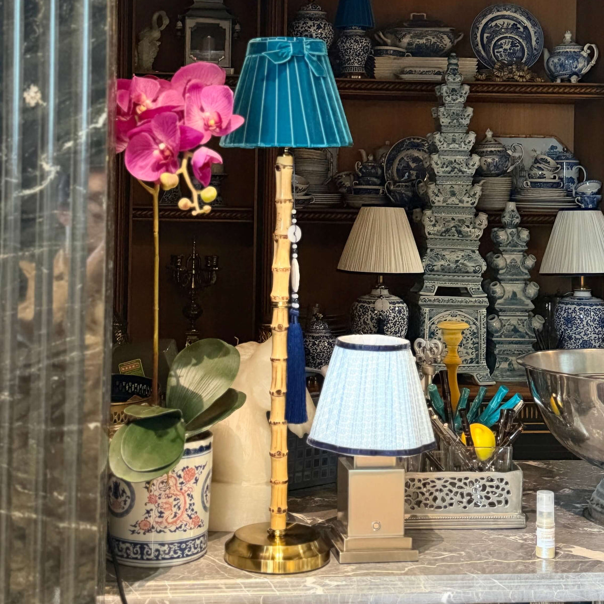 A styled interior scene showing a decorative lamp with a turquoise-blue pleated shade and gold-toned base on a marble surface, alongside other home decor items, ceramic vessels, and a pink orchid in a shop or room setting.