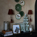A styled interior scene showing a dark wooden dresser against a textured beige wall, decorated with bird-themed ceramic plates, two red lampshades, books, a framed print, and a decorative green object.