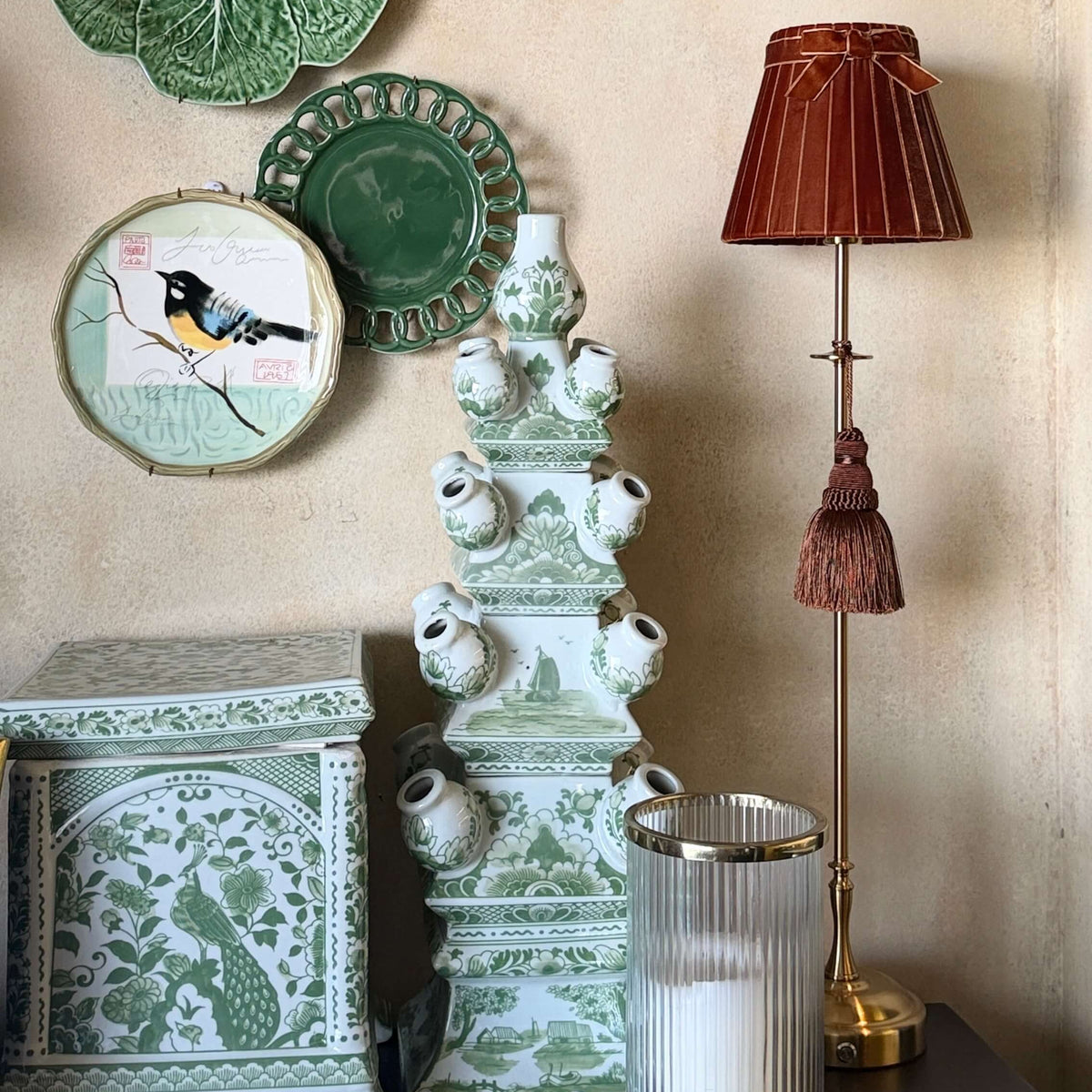 A styled interior scene with decorative ceramic wall plates, a green-and-white patterned chest, a tall sculptural vase, a lit table lamp with a brown pleated shade, and a candle holder against a warm beige wall.