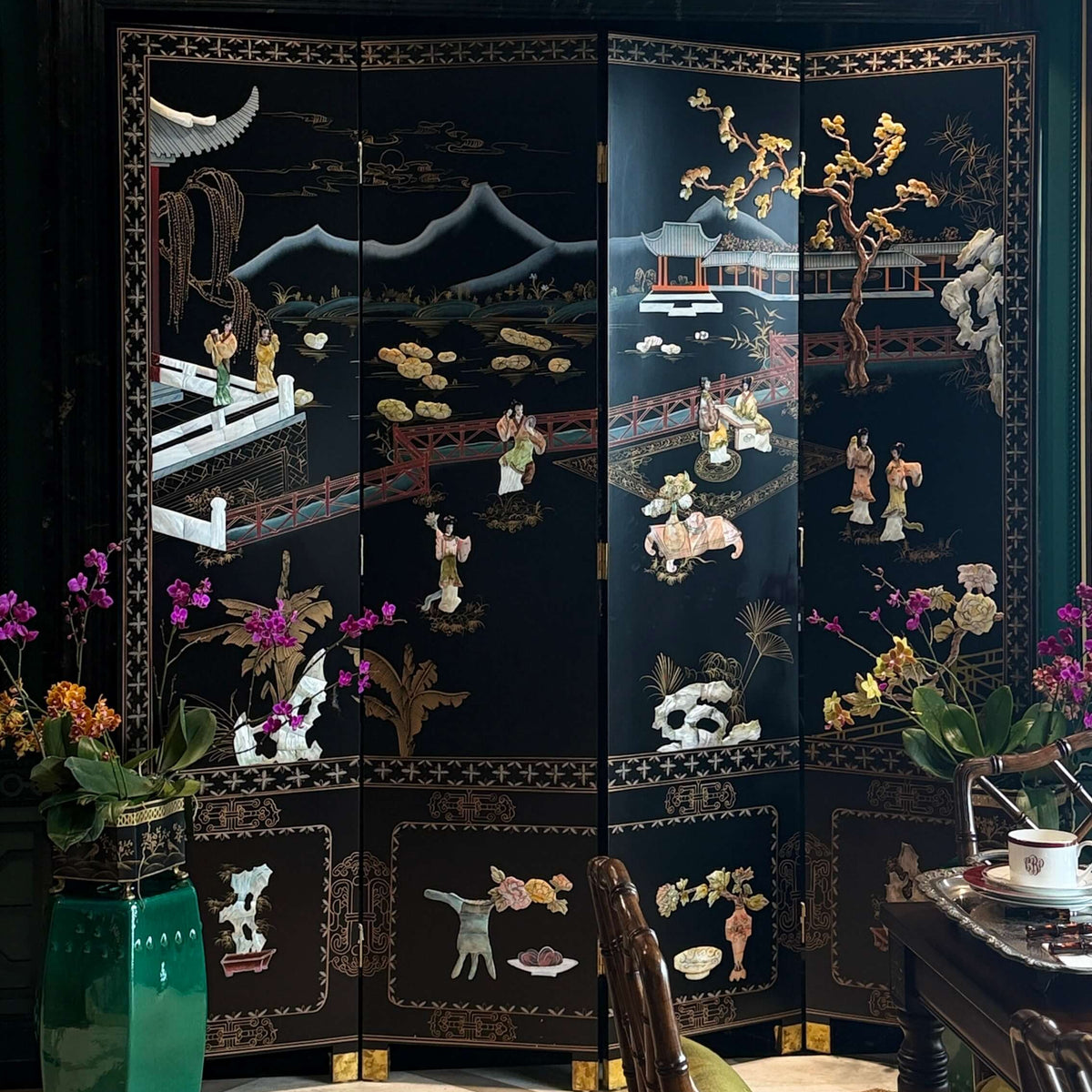 A black lacquered folding screen with intricate mother-of-pearl Chinese landscape and figure motifs, set in a decorated room with a green wall, beige carpet, and floral arrangements.
