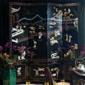 A black lacquered folding screen with intricate mother-of-pearl Chinese landscape and figure motifs, set in a decorated room with a green wall, beige carpet, and floral arrangements.
