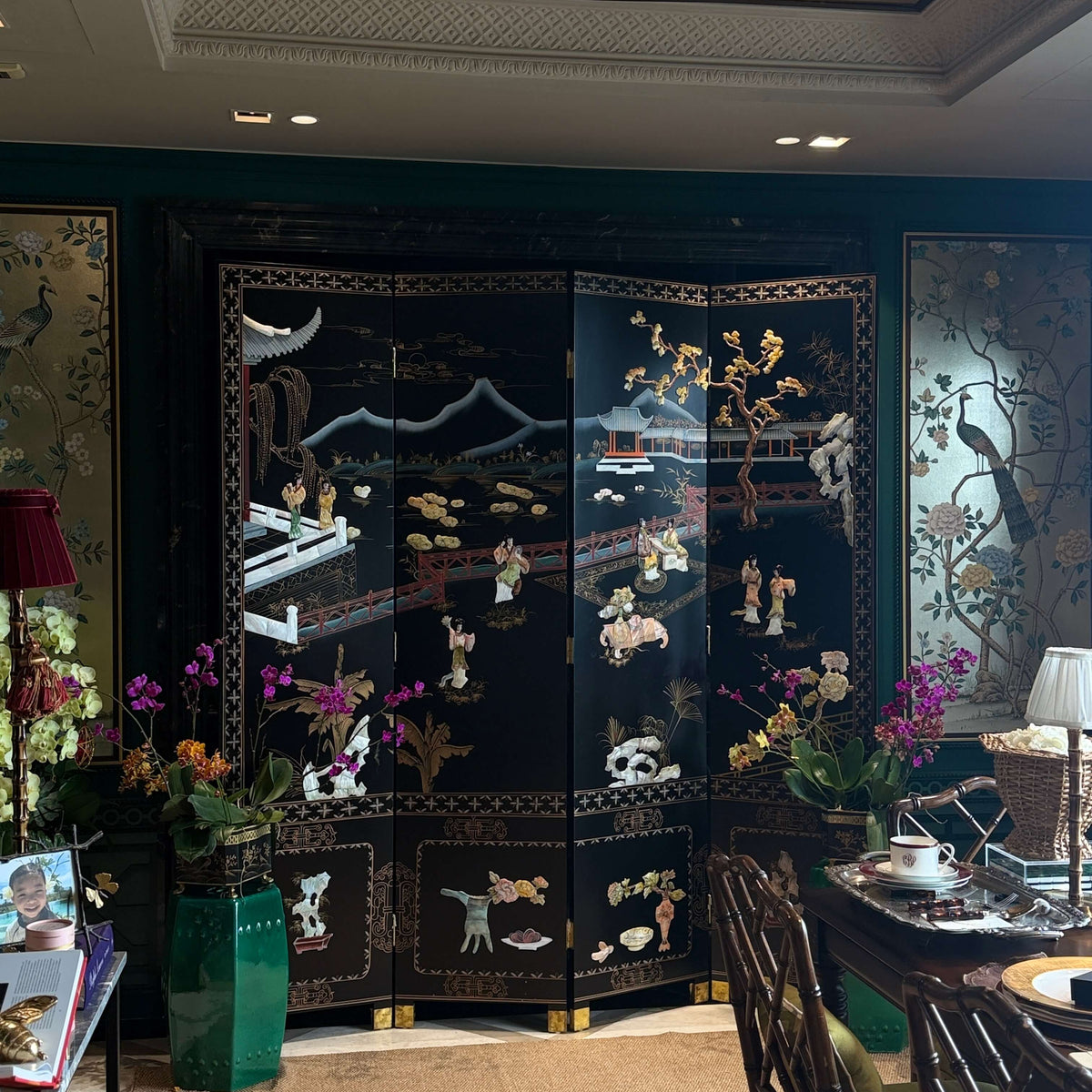 A black lacquered folding screen with intricate gold and multicolor hand-painted Chinese motifs, set in a decorated dining room with green walls and traditional furniture.