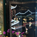 A black lacquered folding screen with intricate hand-painted and inlaid scenes of figures, mountains, boats, and water lilies, styled in a room next to a green glass vase with flowers.