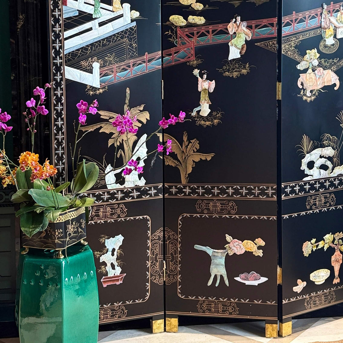 A black folding screen with intricate gold and multicolor mother-of-pearl Chinese landscape and figure illustrations, styled in an interior setting next to a green glass vase with flowers.