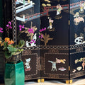 A black folding screen with intricate gold and multicolor mother-of-pearl Chinese landscape and figure illustrations, styled in an interior setting next to a green glass vase with flowers.