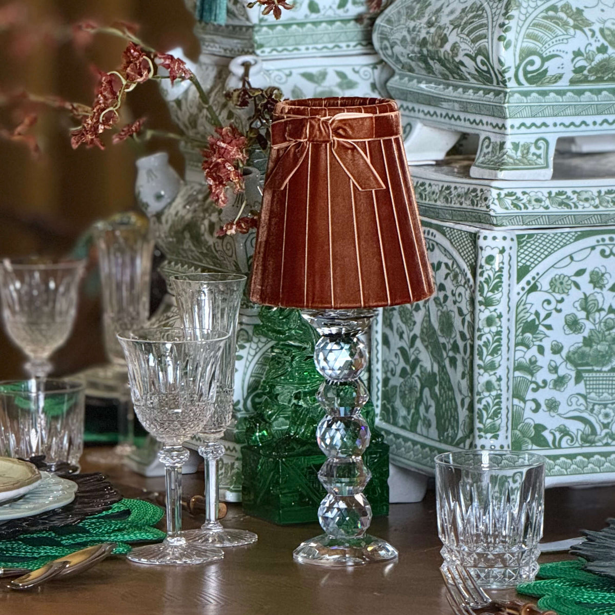 A styled tabletop scene with a small brown pleated table lamp on a clear glass pedestal, surrounded by crystal glasses, green placemats, and ornate green-and-white decorative boxes and floral arrangements.