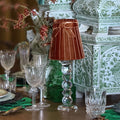 A styled tabletop scene with a small brown pleated table lamp on a clear glass pedestal, surrounded by crystal glasses, green placemats, and ornate green-and-white decorative boxes and floral arrangements.